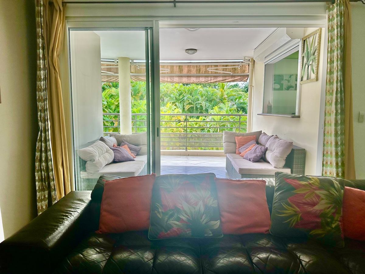 A living area features a spacious dark leather couch adorned with tropical-themed cushions. Large sliding glass doors open to a furnished terrace, showcasing lush greenery outside. Natural light illuminates the space, complemented by light curtains that frame the view.
