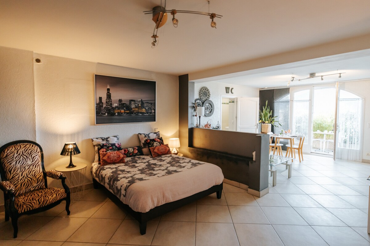 A spacious sleeping area features a large bed adorned with decorative pillows. A variety of lighting options, including a bedside lamp, creates a cozy ambiance. The open layout connects to a well-equipped kitchen area, while natural light floods in from the adjoining sunroom.