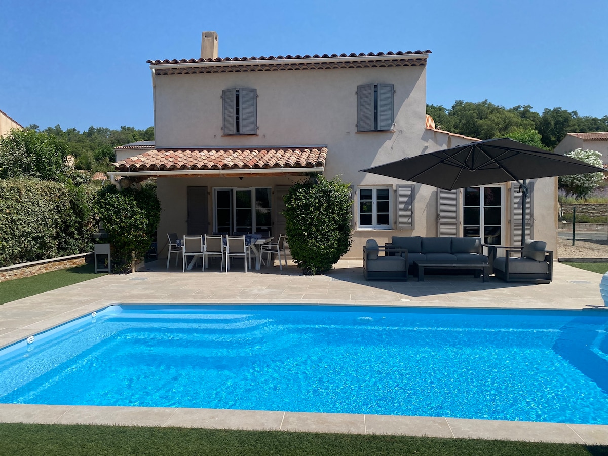 A villa is depicted, featuring a crystal-clear swimming pool in the foreground. The outdoor seating area includes a shaded lounge setup and a dining table. The house has traditional shutters, with a sloping roof, surrounded by lush greenery and a clear blue sky.