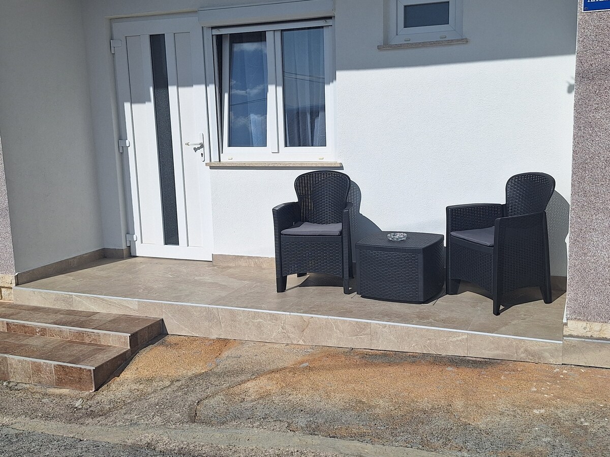 A covered terrace is presented with two black wicker chairs and a small table, providing a welcoming seating area. The entrance features a door with a glass panel, framed by a white wall and a nearby window, allowing for natural light.