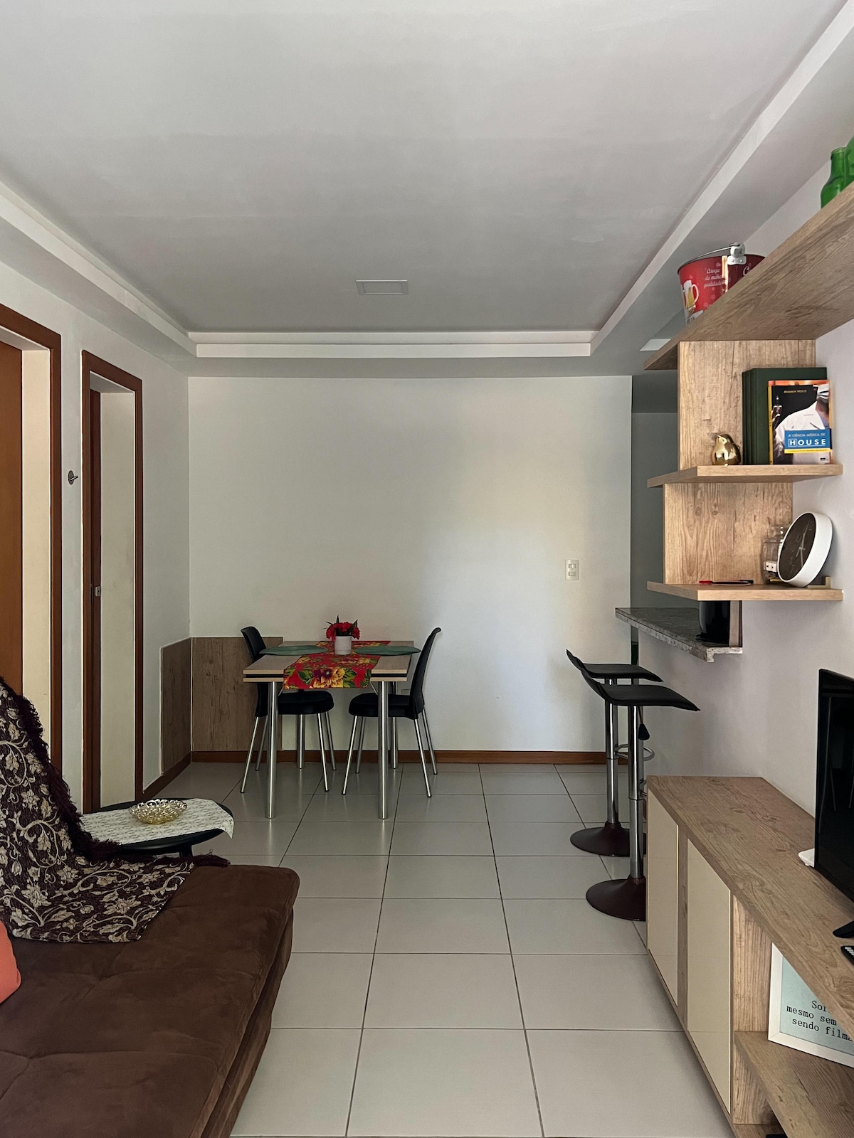 A small living area is presented, featuring a compact dining table with four chairs. Adjacent to the table, two bar stools are positioned by a countertop. A neutral palette is complemented by natural light filtering through a window, highlighting the modern furniture and decor.