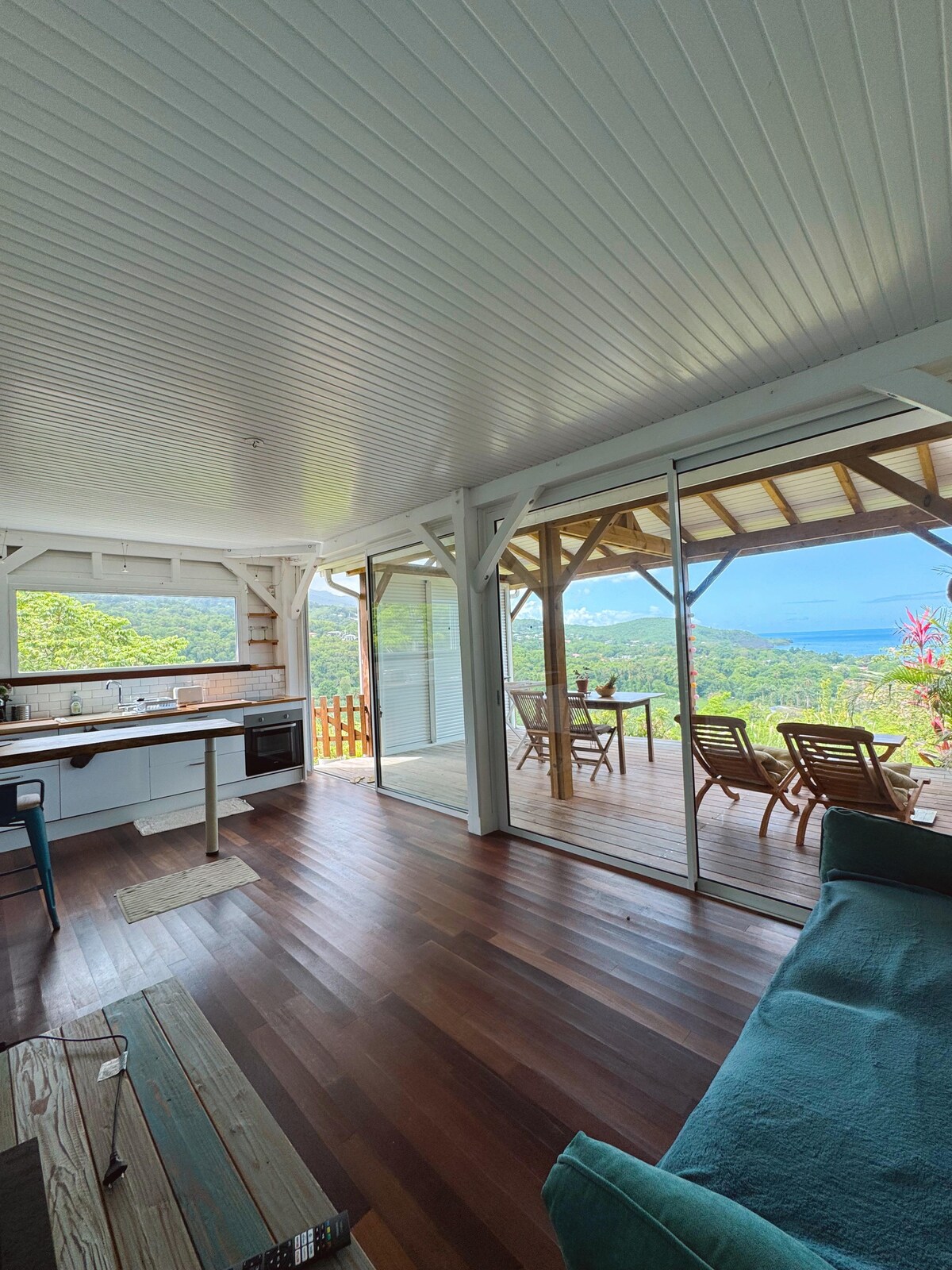 A spacious living area features wooden flooring and a well-equipped kitchen. Floor-to-ceiling glass doors open to a deck with stunning views of the surrounding mountains and the Caribbean Sea. Relaxing seating options are present, providing a comfortable atmosphere.
