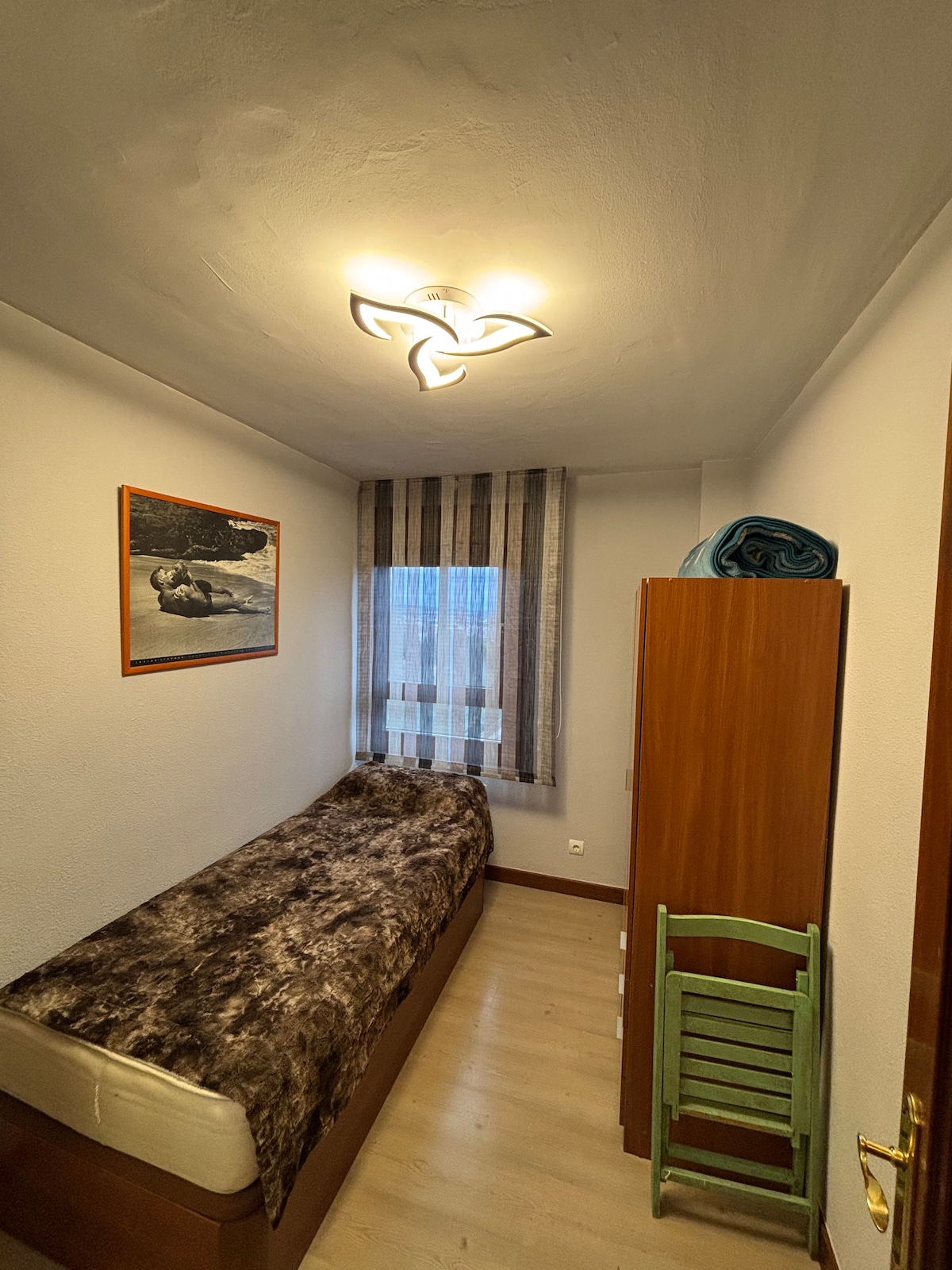 A compact sleeping area is furnished with a single bed featuring a patterned bedspread. A wardrobe is positioned against the wall, complemented by a green chair. Natural light enters through a window with striped curtains, and a decorative wall art piece adds a subtle touch.