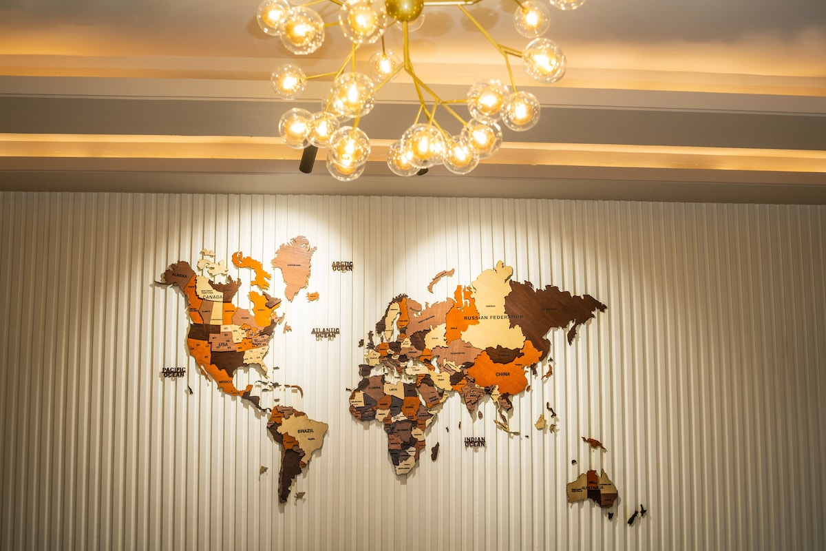 A wooden world map adorned with various shades of orange and brown is prominently displayed on a textured wall. Above, a modern chandelier with multiple bulbs adds an elegant touch to the room, providing a stylish focal point.