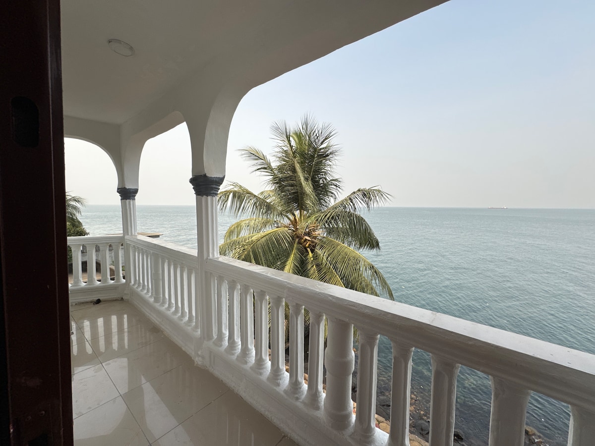 A spacious balcony features classic white railings, overlooking calm waters and lush palm trees swaying gently in the breeze. The serene view offers a sense of tranquility, with a clear horizon extending into the sky.