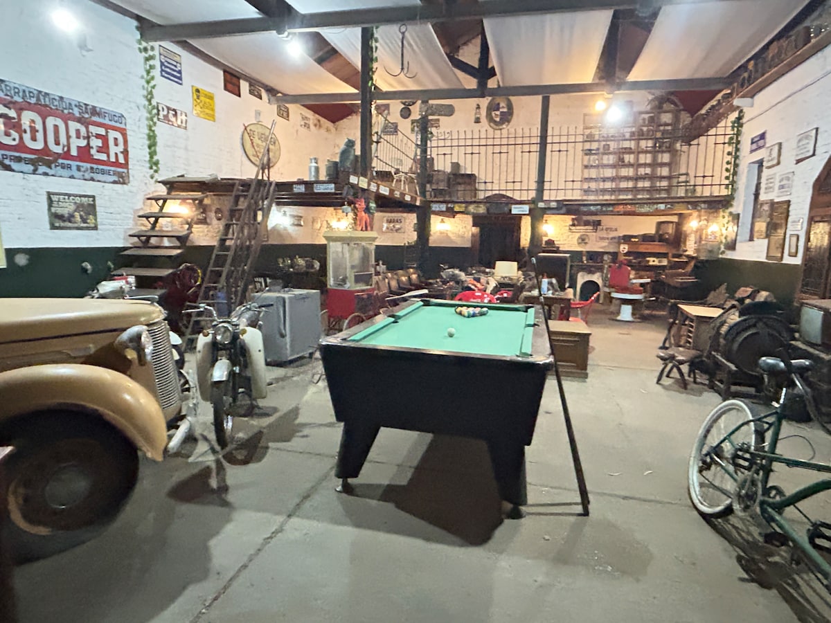 A spacious interior features vintage decor including various antiques and collectibles. A billiard table occupies the center, surrounded by old vehicles and furniture. Historical signage adorns the walls, adding character to the space. Warm lighting enhances the unique ambiance created by the eclectic items.