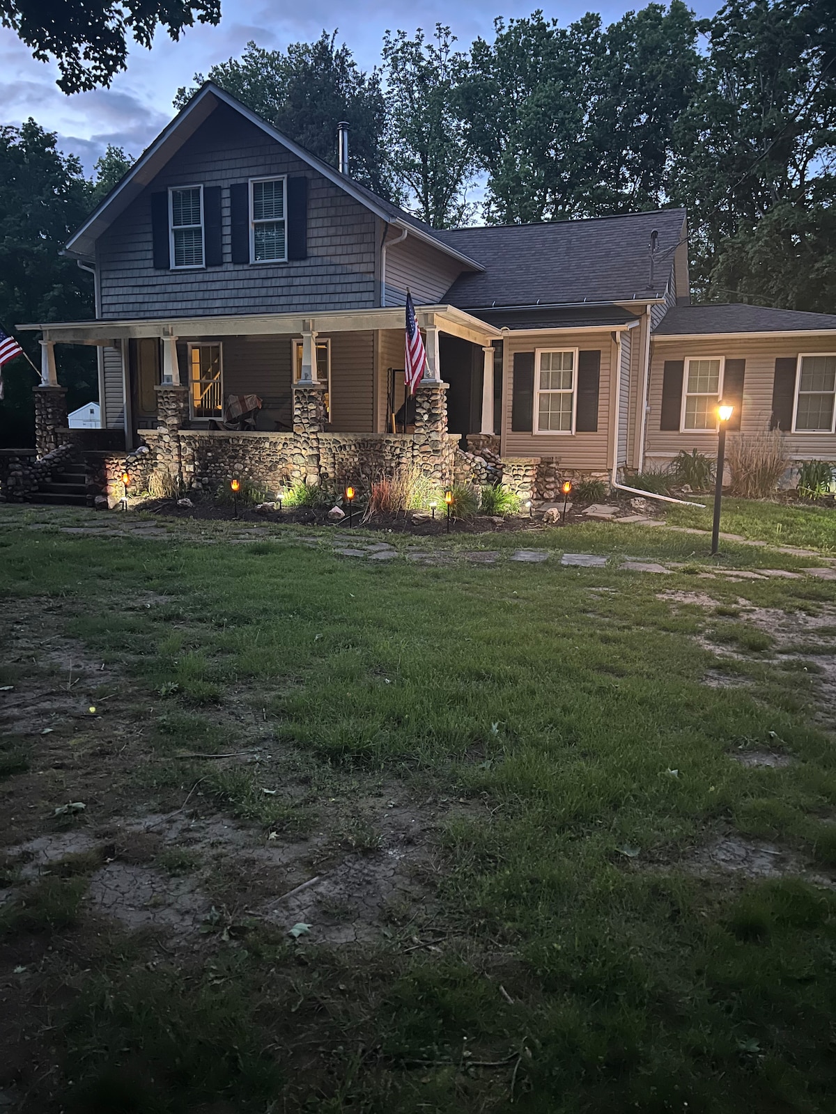 The front view of a renovated craftsman home is displayed, featuring a welcoming porch with flagstone accents and American flags. Soft garden lighting highlights the landscape, which includes a well-maintained lawn and stone pathways leading up to the entrance.