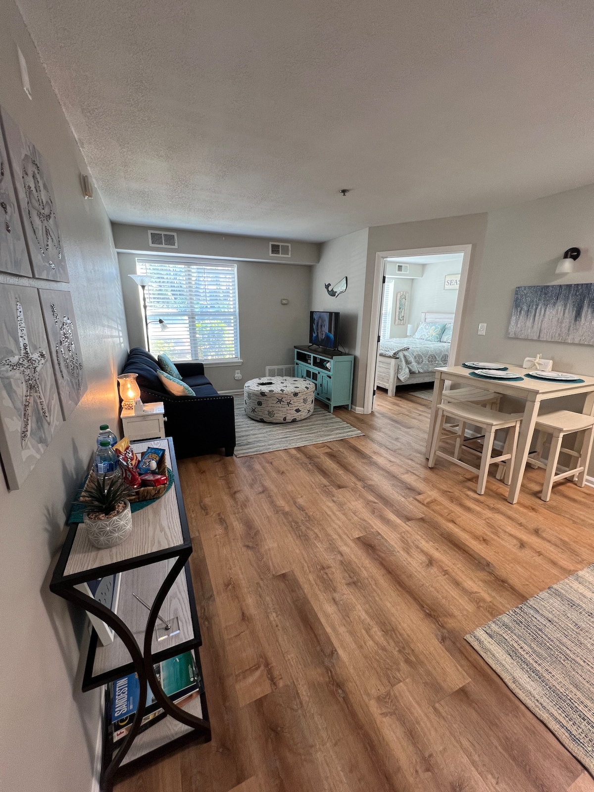 A cozy living area is visible, featuring a dark sofa and decorative pouf. Natural light enters through multiple windows, illuminating the wood-style flooring. A small dining table is positioned nearby, and a corner entertainment unit holds a television. Art is displayed on the wall, enhancing the space.