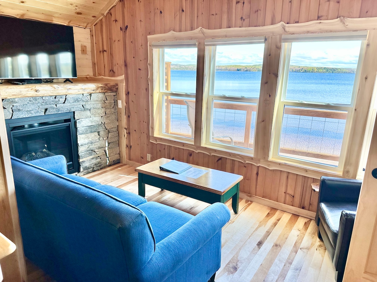 A living area features a cozy blue sofa and a black chair, positioned near a stone fireplace. A coffee table sits in front of the sofa, while three large windows offer an expansive view of AuTrain Lake, illuminating the space with natural light.