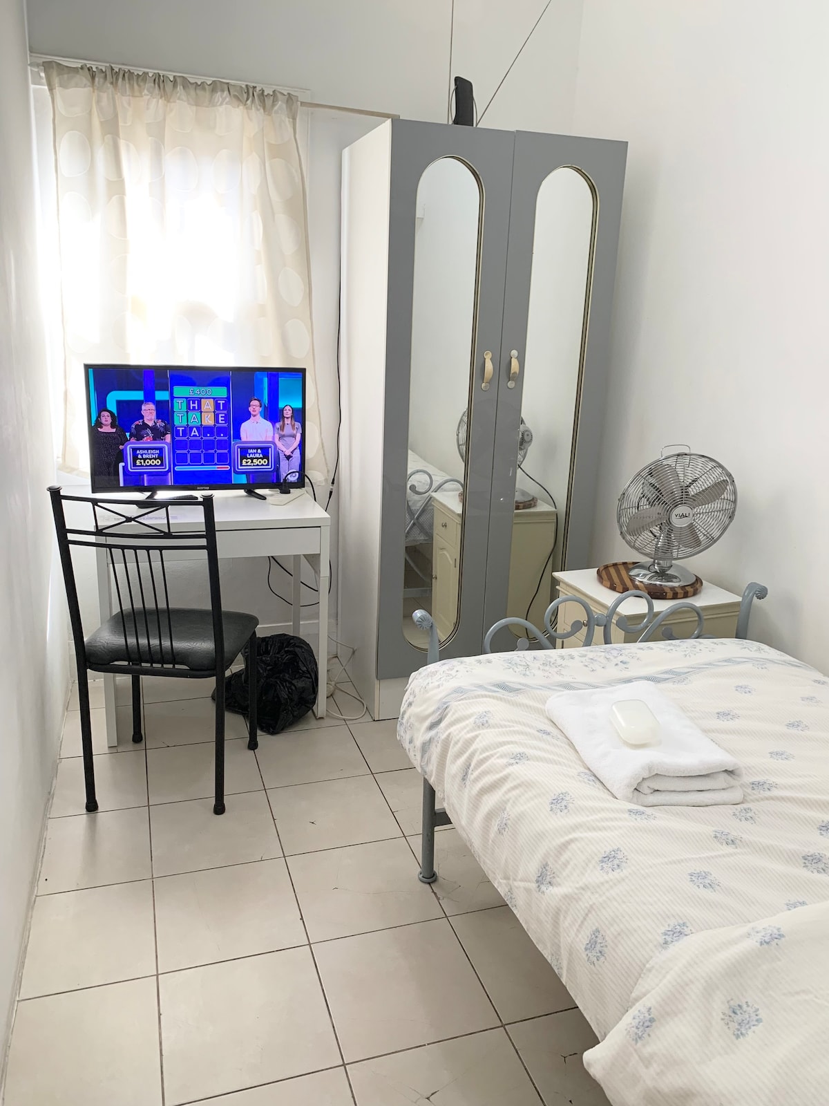 A compact room features a simple bed with light bedding, a television mounted on a white desk, and a chair. A wardrobe with mirrored doors stands beside the bed, while a fan offers additional comfort. Natural light filters through sheer curtains, enhancing the space.