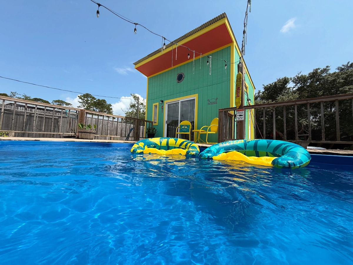 A vibrant green structure is visible in the background, complemented by yellow and orange accents. In the foreground, a clear blue pool reflects the sky, with two colorful inflatable loungers floating on the surface. String lights hang above, suggesting a lively atmosphere.