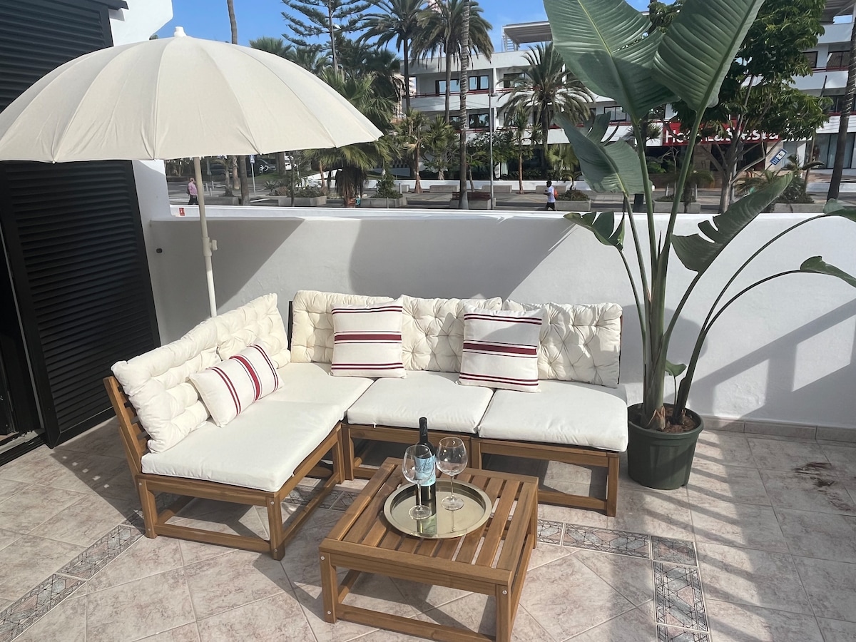 A relaxing terrace area features a sectional sofa with soft cushions and accent pillows, alongside a wooden coffee table. An umbrella provides shade, enhancing comfort. A potted plant adds a touch of greenery, while palm trees are visible in the background.