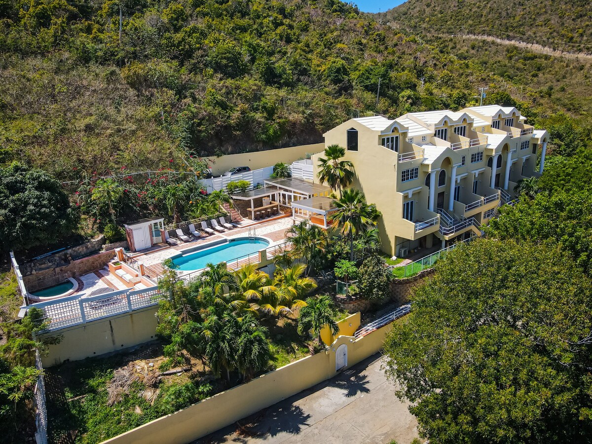 An aerial view of a welcoming multi-unit structure surrounded by lush greenery. A shared pool is visible in the foreground, accompanied by lounge chairs and palm trees. The vibrant yellow building harmonizes with the tropical landscape, creating an inviting space for relaxation.