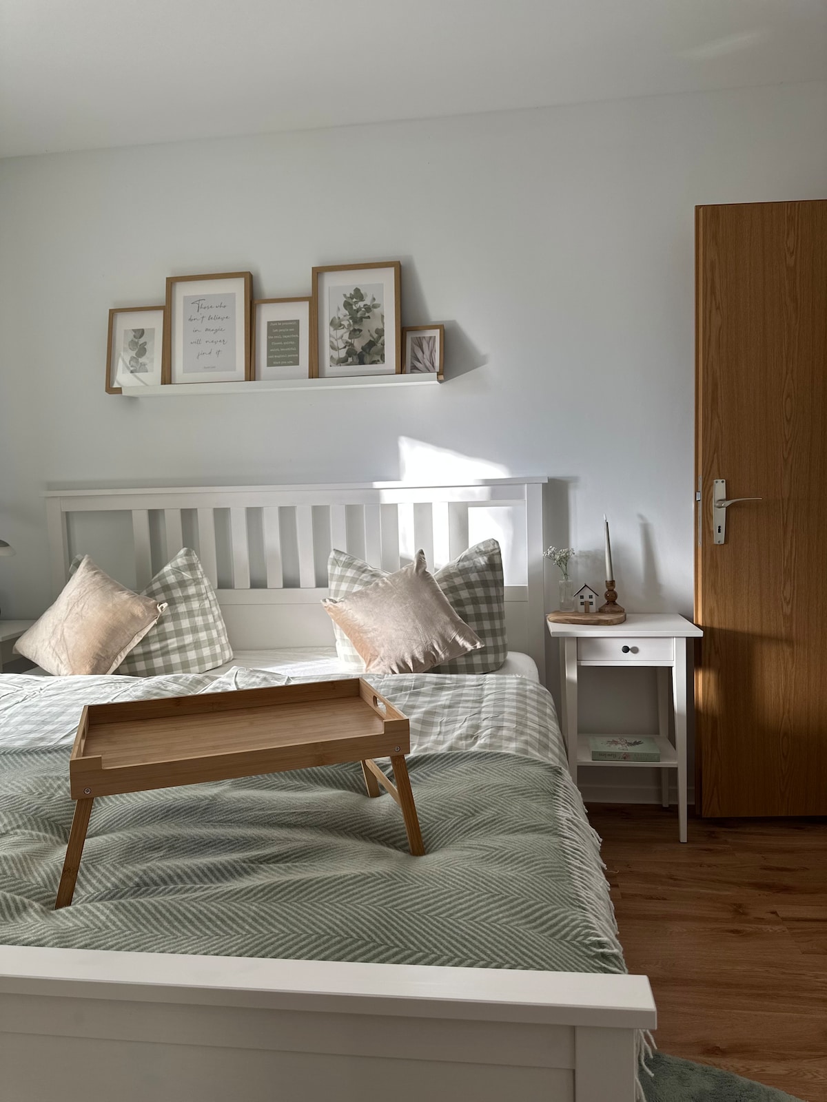 A bright bedroom features a spacious bed with decorative pillows and a cozy blanket. A wooden tray rests atop the bed, while a bedside table holds a small lamp. Light walls and natural light enhance the room's calming atmosphere.