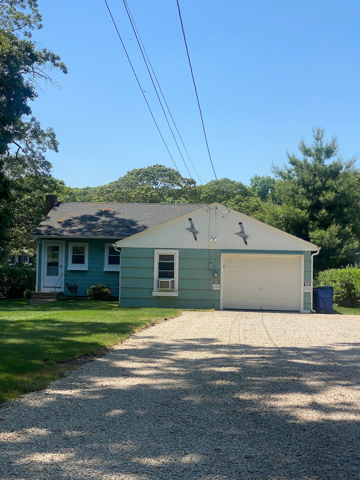The cottage features a light blue exterior with a single-car garage and a front door framed by a small porch. The gravel driveway provides access to the home, surrounded by well-kept greenery and trees, enhancing the quiet neighborhood ambiance.
