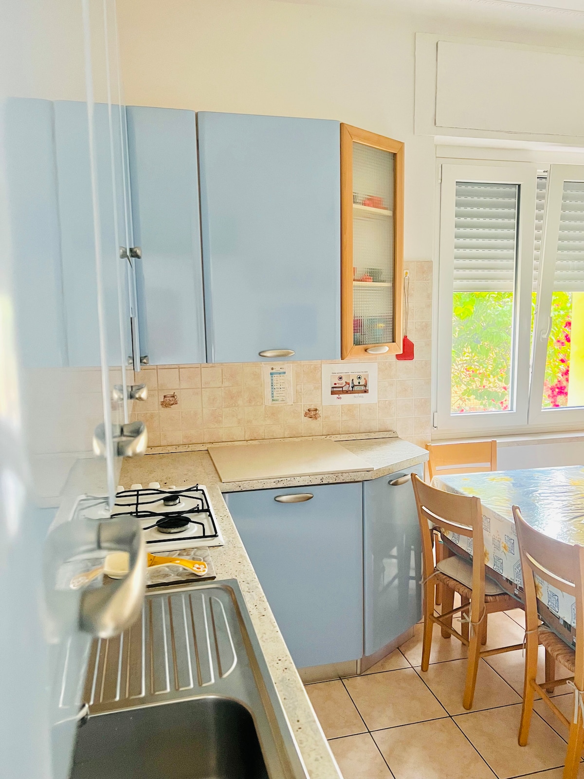 The kitchen area is designed with light blue cabinetry, offering a bright and airy feel. Countertops are made of a light stone material, and a stainless steel sink is visible. A dining table with light-colored chairs is set near a window that allows natural light to enhance the space.