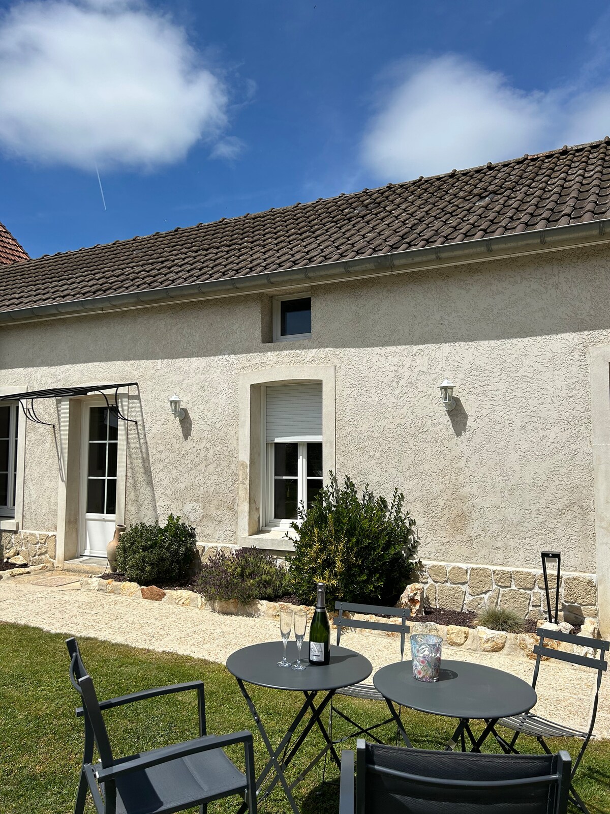 An outdoor seating area features a small round table set with two flutes and a bottle of champagne. Gray chairs are positioned around the table, with a garden visible beyond. A light-colored stone facade is paired with a slate roof, while a few windows add to the charm.