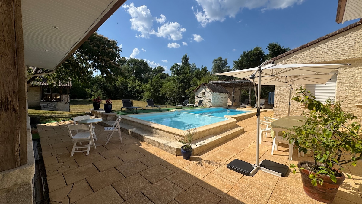 An outdoor area features a clear swimming pool surrounded by natural stone, with patio tiles laid across the space. White lounge chairs and tables are set under an umbrella, with potted plants adding greenery. Lush trees and open fields can be seen in the background.