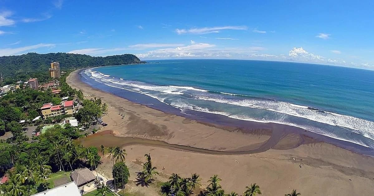 An aerial view captures the contrast of sandy beach meeting blue ocean waters, with gentle waves rolling onto the shore. Lush green hills are visible in the background, accompanied by scattered trees along the coastline and buildings that reflect a tranquil coastal environment.