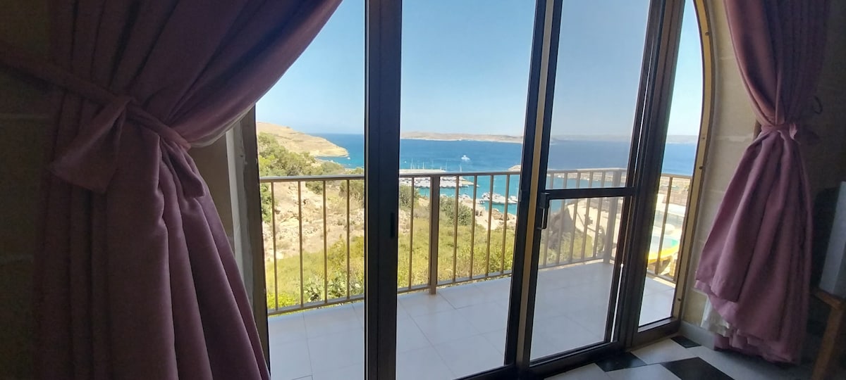 A balcony door framed by soft pink curtains provides access to stunning views of the Gozo channel and the surrounding landscape. Bright sunlight reflects off the serene water, creating a calm atmosphere. The tiled floor inside complements the natural scenery visible through the large glass panels.