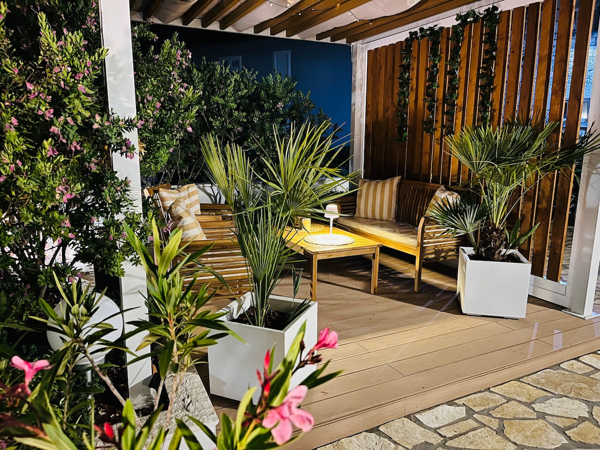 A seating area is surrounded by lush greenery and flowers. Wooden furniture provides a relaxed atmosphere, accompanied by potted plants in white containers. The structure features wooden slats overhead, creating a cozy and inviting space for guests to enjoy the outdoors.