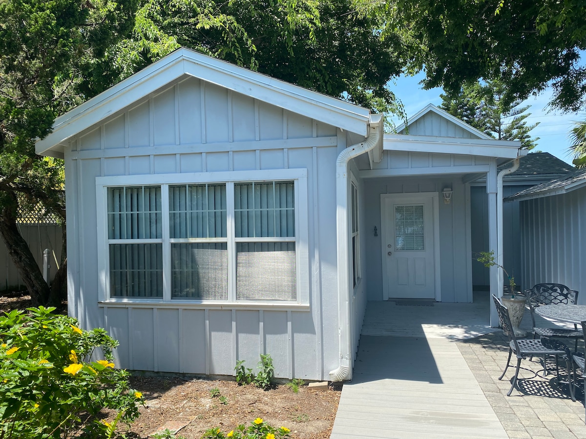 The exterior of the cottage showcases a light-colored facade with large front windows, allowing ample natural light. A welcoming entryway leads to a small patio space furnished with a table and chairs. Lush greenery surrounds the entrance, enhancing the serene environment.