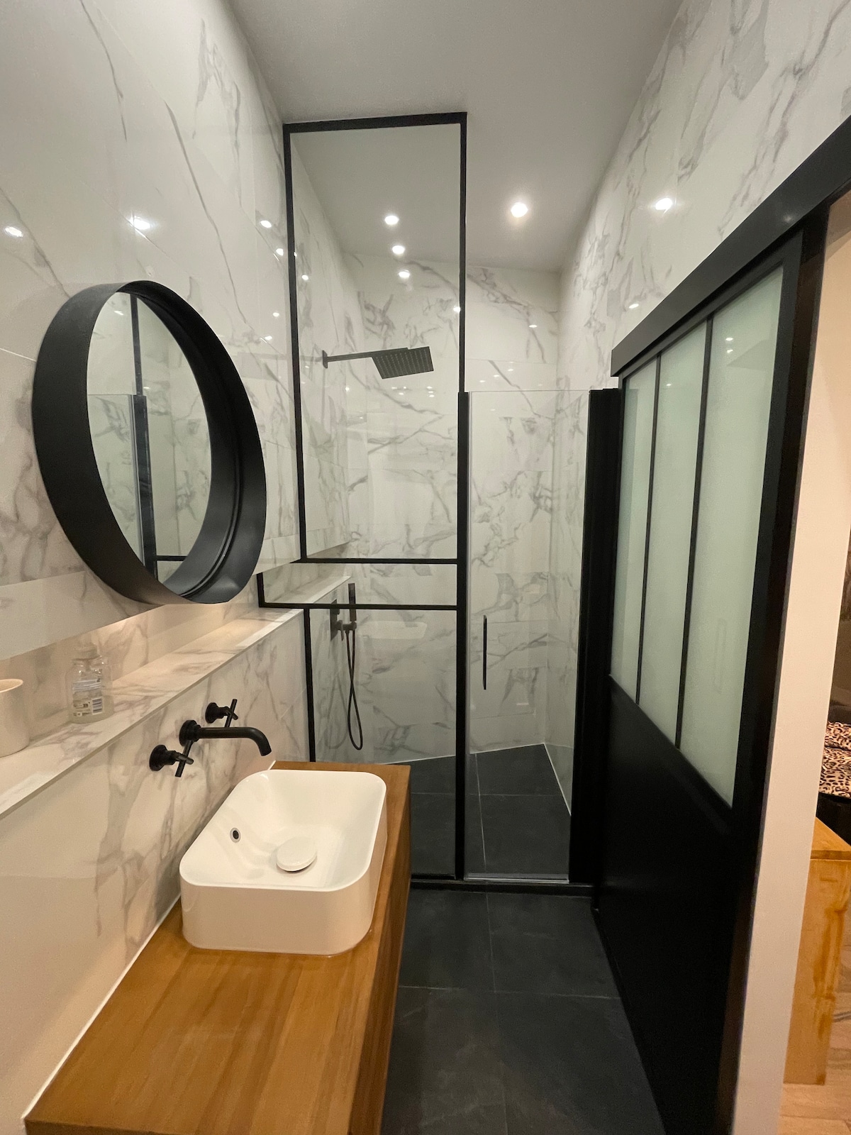 The bathroom features modern fixtures, including a sleek round mirror and a minimalist sink atop a wooden vanity. A frameless glass shower enclosure showcases elegant marble walls, while stylish black accents are present in the hardware and sliding door.