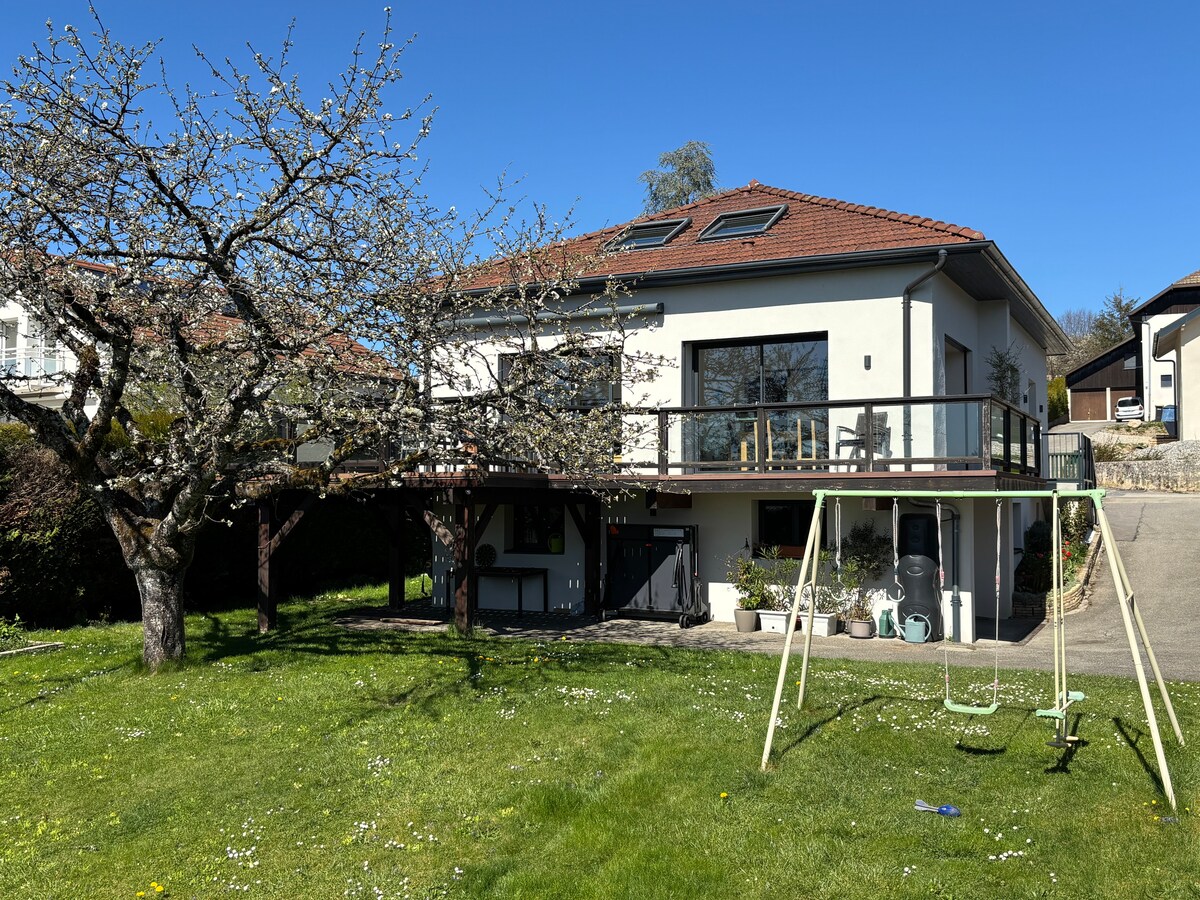 The modern house features a spacious upper deck with glass railings, surrounded by a lush garden. A swing set is visible in the foreground, under the shade of a blooming tree. Large windows provide a view of the backyard and flood the interior with natural light.