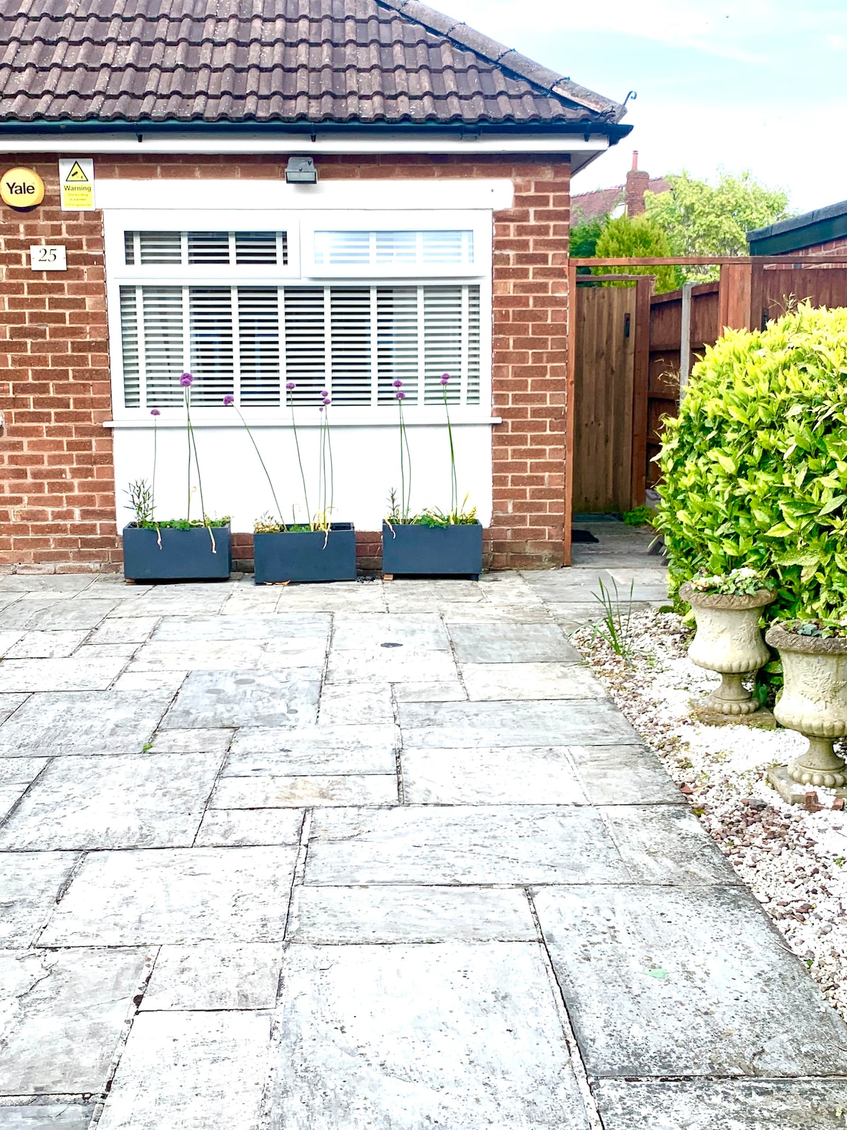 The exterior of the bungalow is showcased, featuring a brick façade with a large window adorned by white shutters. Planters with tall flowers are arranged in front, complemented by a paved patio area and neatly trimmed hedges, creating a welcoming entrance.