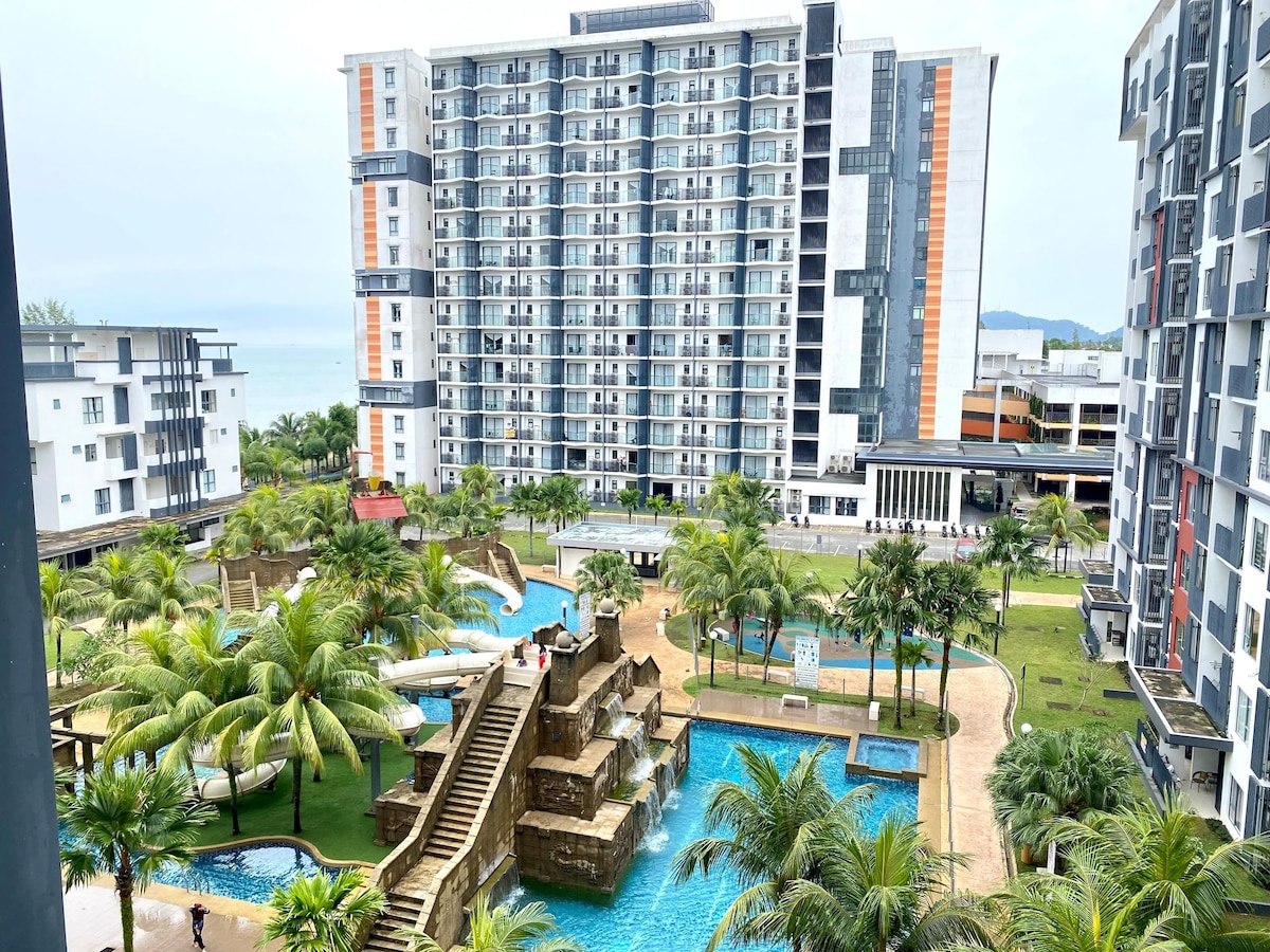 A resort complex is depicted, featuring multiple modern buildings surrounding a water park area. Several pools are visible, interspersed with tropical palm trees and landscaped greenery. The pathways lead to various levels with bridges connecting the different sections, offering a serene environment near the beach.