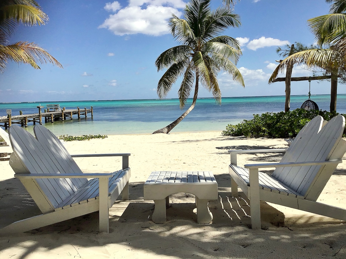 Two white Adirondack chairs are positioned on the sandy beach, facing the calm waters of the Caribbean. A small table stands between the chairs, surrounded by palm trees. In the background, a wooden dock extends into the turquoise sea.