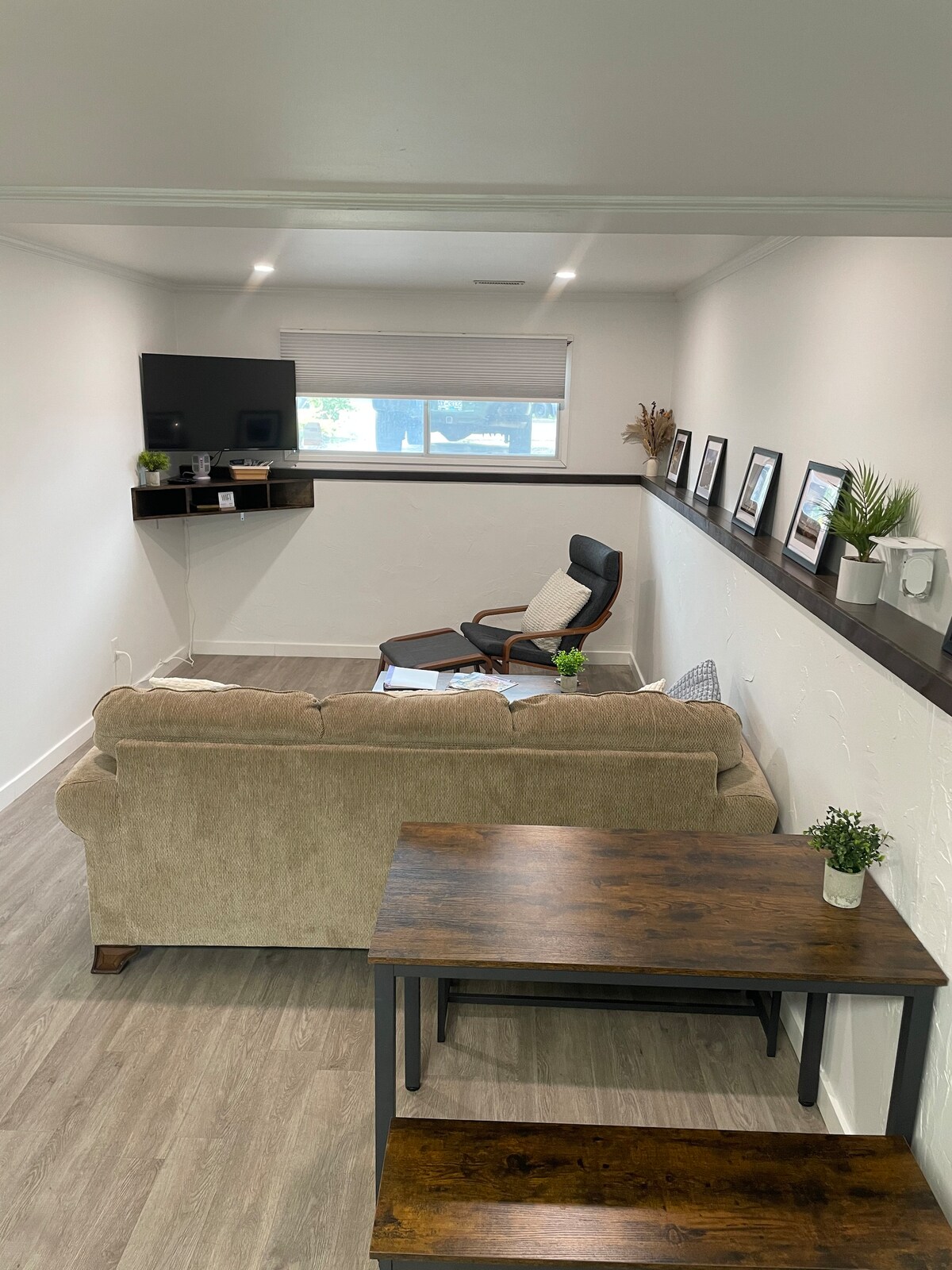The living area features a comfortable sofa facing a wall-mounted television. A cozy armchair is positioned nearby, and a wooden coffee table complements the space. Framed photographs and small plants adorn the shelf above, enhancing the modern decor.
