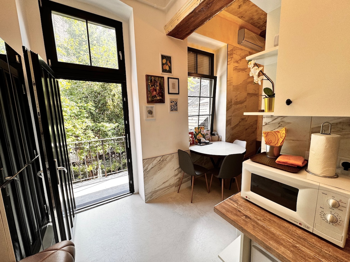 A modern kitchen area features a microwave and a small dining table with four chairs, positioned near the window. Natural light filters in through the glass doors, which open to a balcony overlooking greenery, creating a bright and functional space.