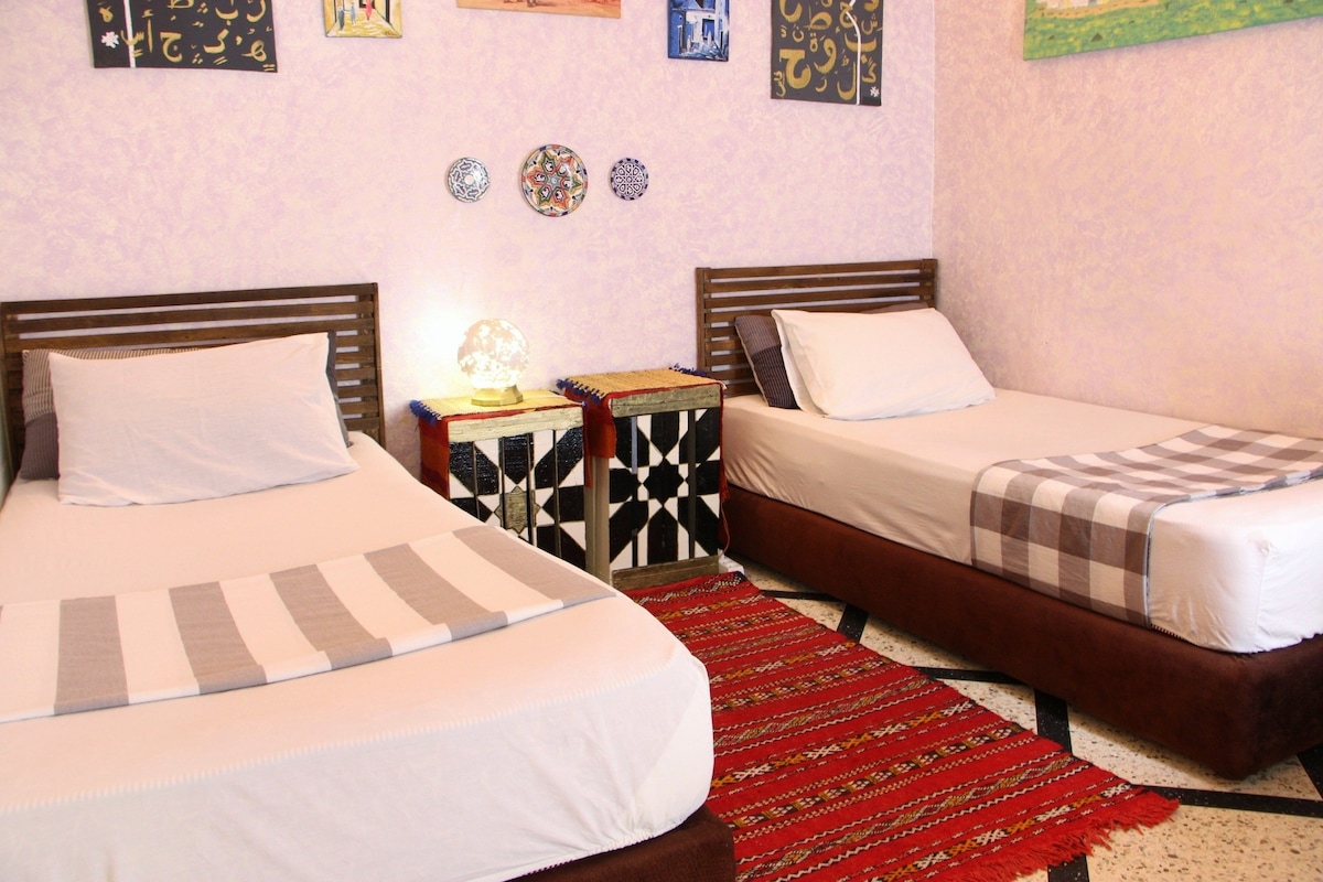 The room features two spacious beds positioned beside intricately designed bedside tables. Colorful artwork adorns the walls, contributing to the inviting ambiance. A woven rug lies on the floor, adding texture to the space.