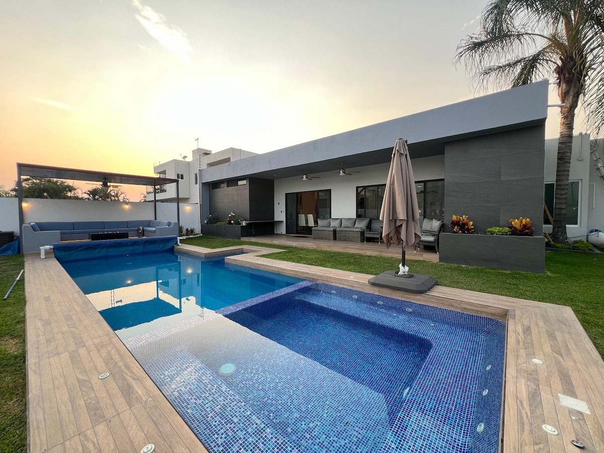 The exterior view showcases a modern one-story house with a spacious patio. A heated pool is surrounded by a wooden deck and lush greenery. Comfortable seating areas are arranged under an umbrella, offering opportunities for relaxation. The scene captures a tranquil sunset in the background.