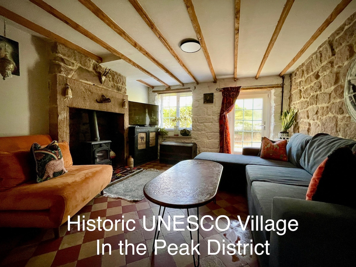 A cozy lounge area features exposed stone walls and wooden beams, providing a warm historical ambiance. A grey sectional sofa and an orange settee are paired with a coffee table, while a television and wood burner add modern comforts. Natural light streams through the window.