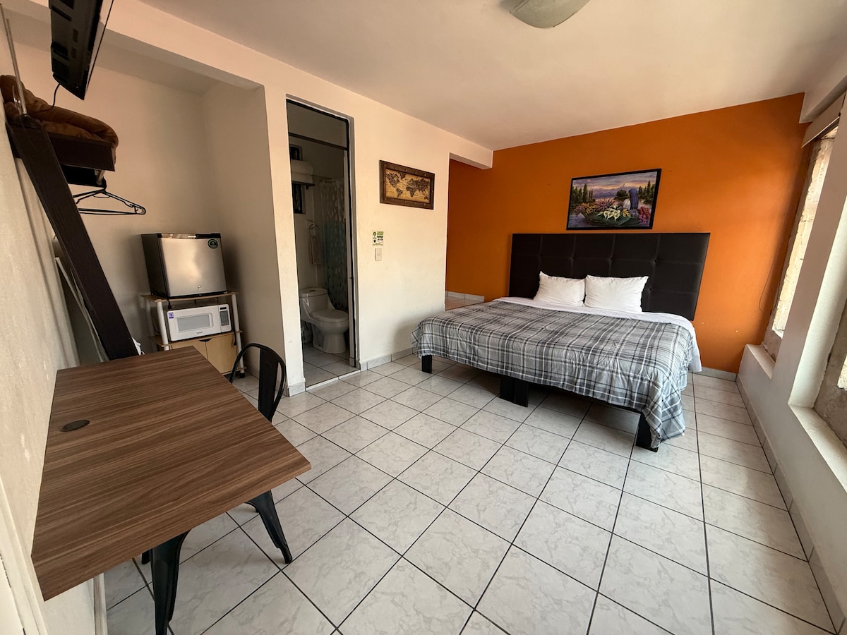 A spacious room features a large bed with a patterned blanket, complemented by two white pillows. A wooden desk and chair are positioned near the window, offering natural light. An orange accent wall and artwork add color, while a bathroom door is visible in the background.