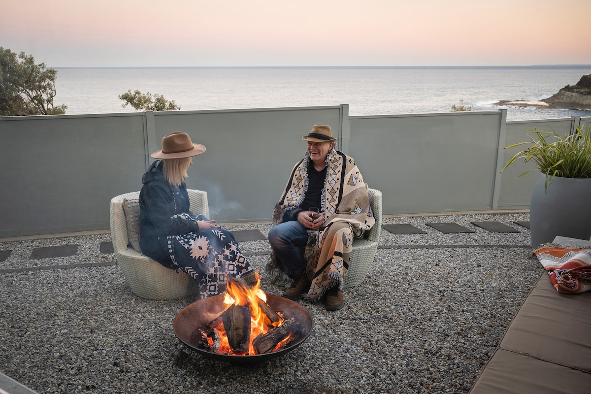 A cozy outdoor seating area features two chairs positioned around a fire pit, with a gentle ocean view in the background. Soft, warm light illuminates the scene as twilight approaches, enhancing the tranquil environment. Decorative blankets are draped over the chairs, adding comfort to the setting.