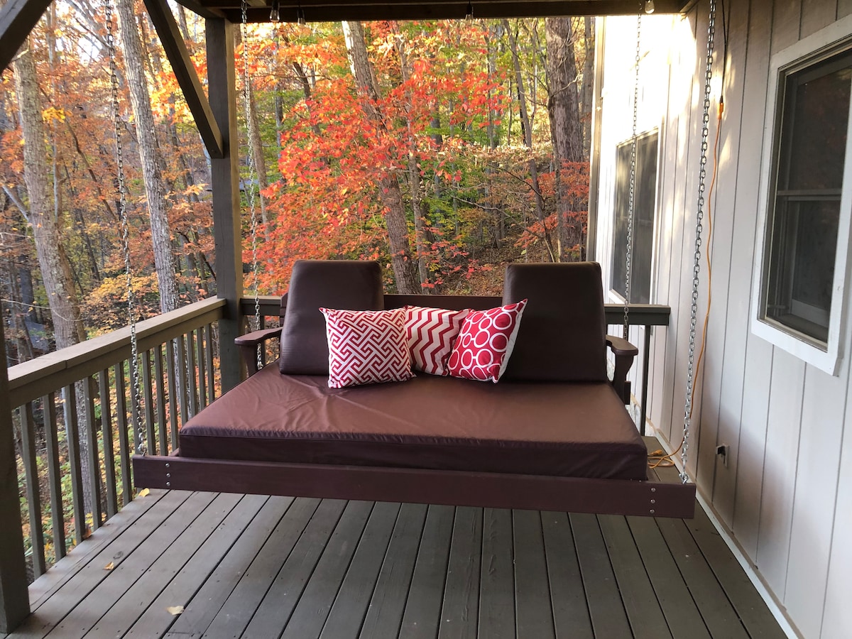 A swing bed with a comfortable cushion is positioned on a private deck, surrounded by trees adorned with autumn foliage. Two decorative pillows add a touch of color. The deck flooring is wooden, contributing to a natural atmosphere.