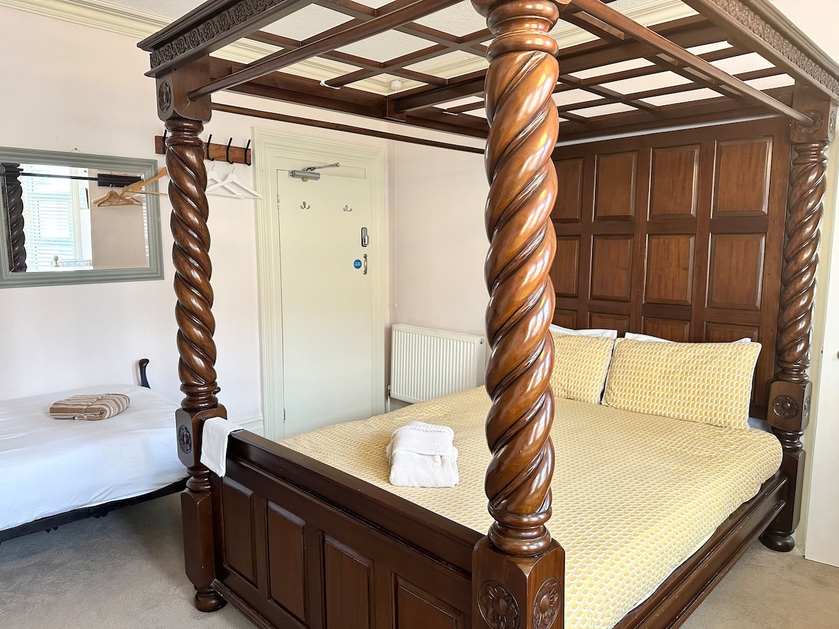 A wooden four-poster bed with intricately carved posts occupies the center of the room, adorned with yellow patterned bedding. A simple white towel is neatly placed on the bed. A mirrored wall adds depth, while a single bed is positioned against the side wall.