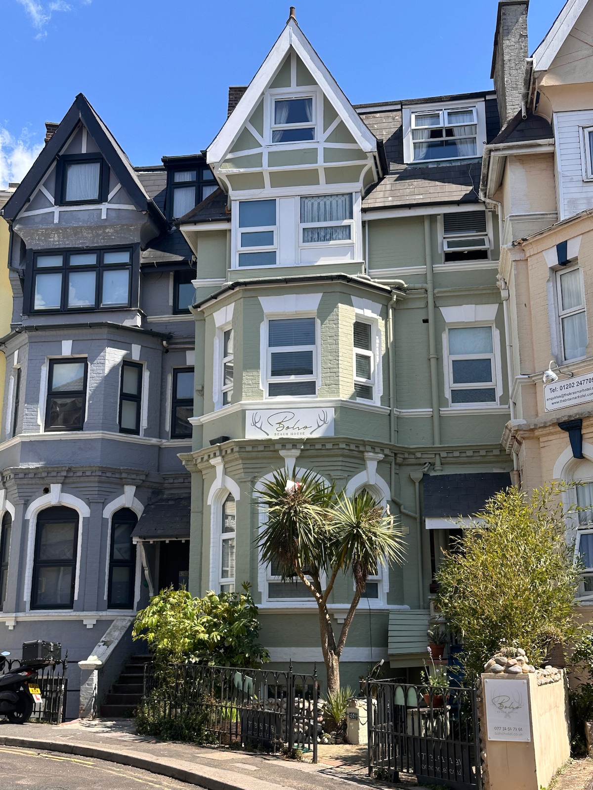 A charming Victorian building is depicted, showcasing a pale green exterior with white trim and intricate architectural details. Large windows and a welcoming entryway are visible, along with palm trees and greenery enhancing the accessible outdoor space.