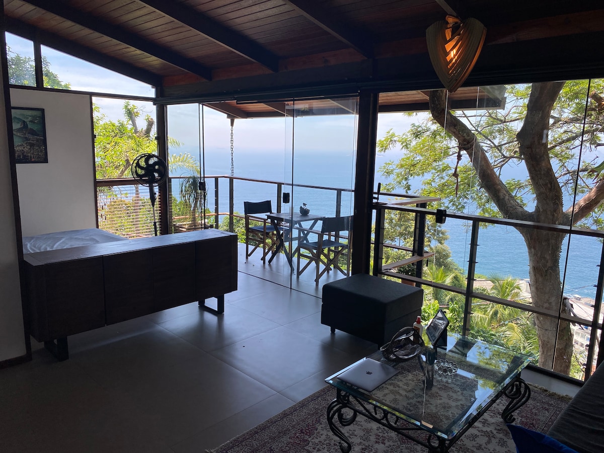 A spacious living area is highlighted with large glass doors, offering unobstructed ocean views. A wooden cabinet is positioned near a comfortable seating area with a low table. Outdoor seating is visible on the balcony, surrounded by lush trees.