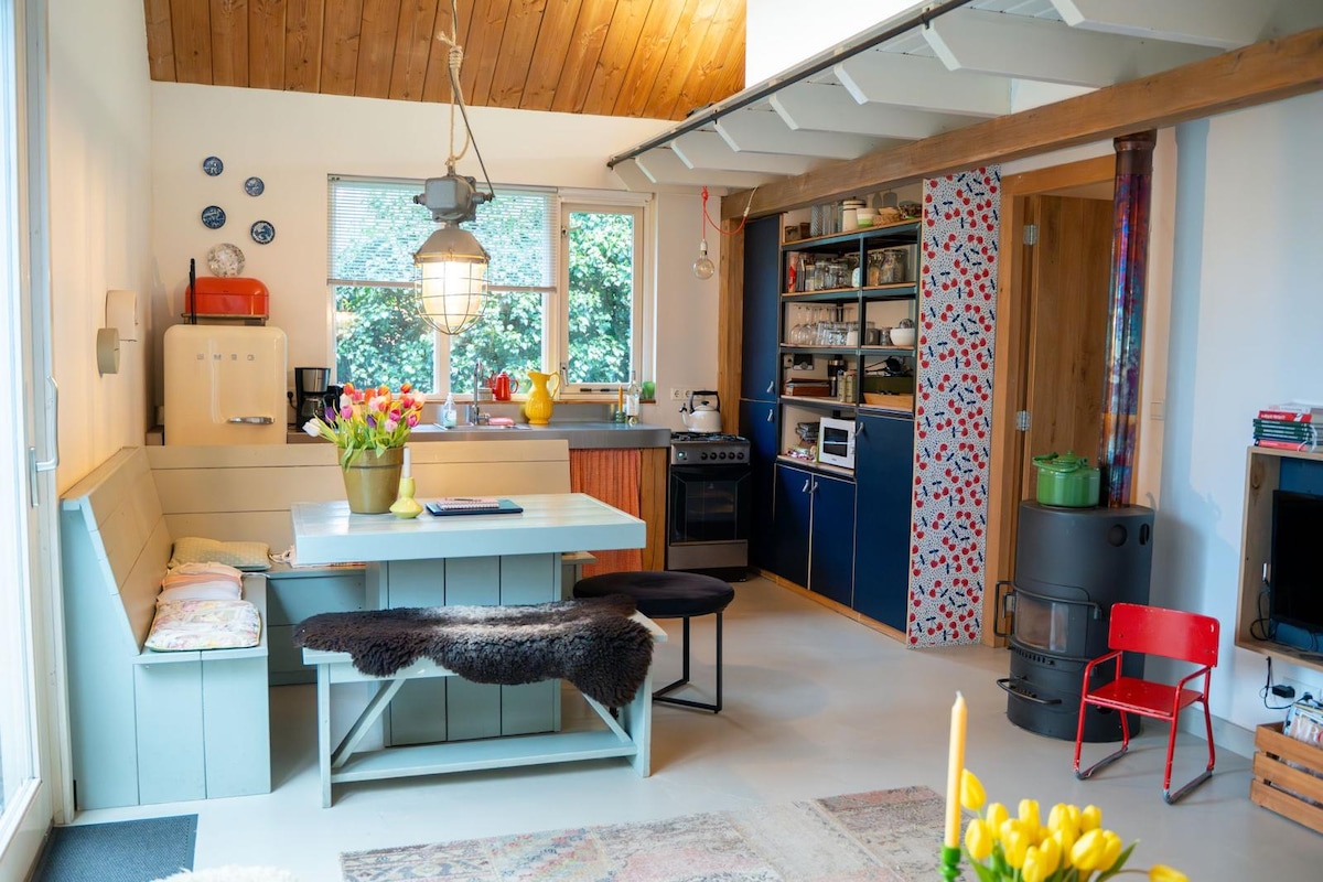 A light-filled kitchen features a cozy dining area with a wooden table and bench, adorned with fresh flowers. The kitchen is equipped with modern appliances, including an oven and refrigerator, set against a backdrop of colorful cabinetry. Natural wood beams add warmth to the space.
