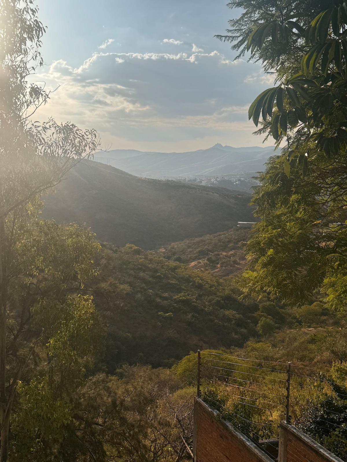 A scenic view of rolling hills and valleys is captured under a soft afternoon light. Lush greenery frames the foreground, leading the eye toward distant mountains. The sky displays a mix of clouds and sunlight, enhancing the natural beauty of the landscape.