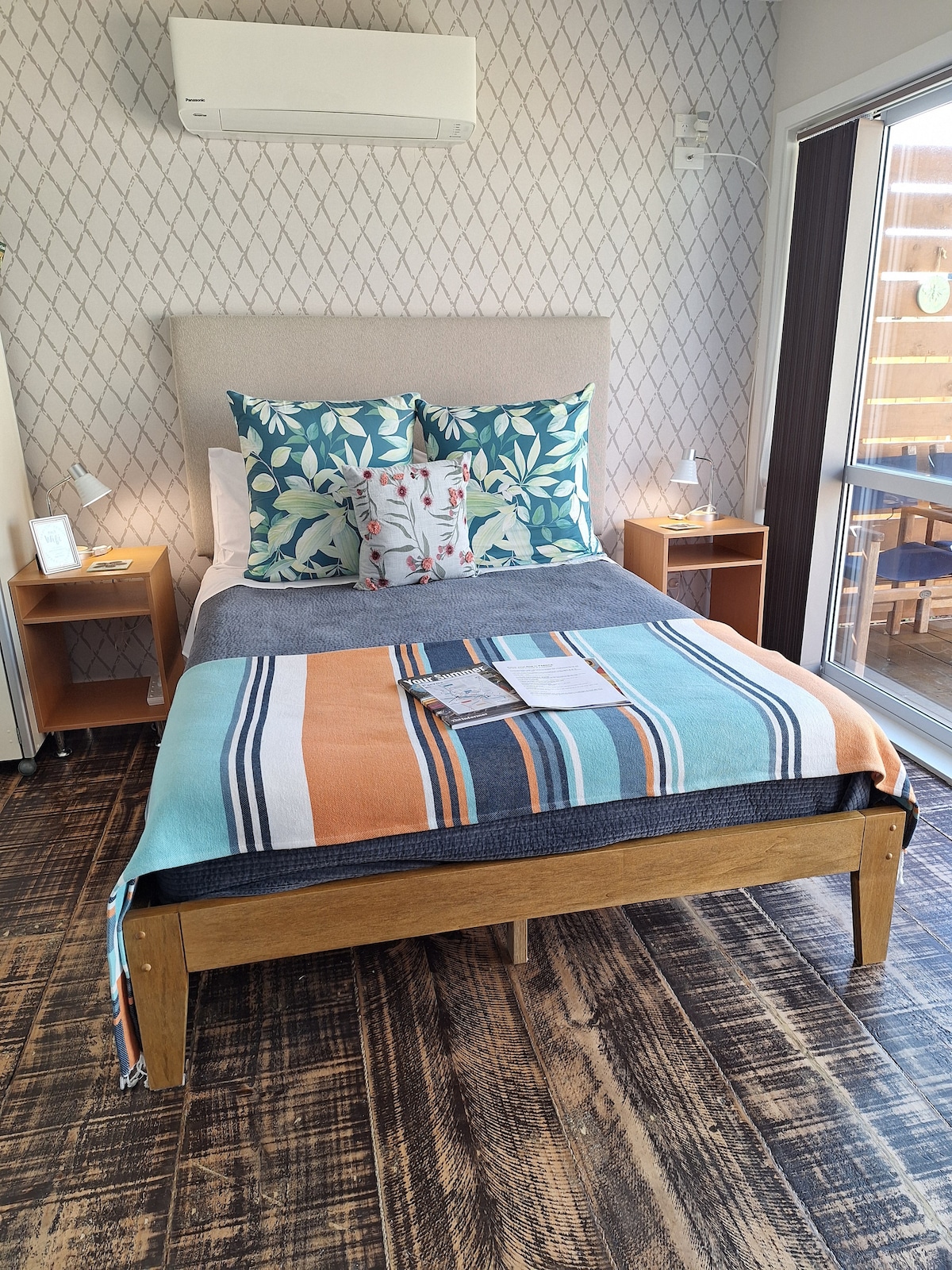 A comfortable bed is positioned in the room, adorned with quality linens and decorative pillows featuring botanical patterns. Soft bedside tables are placed on either side, with lighting fixtures adding functionality. A vibrant blanket drapes across the foot of the bed. Large windows provide natural light.