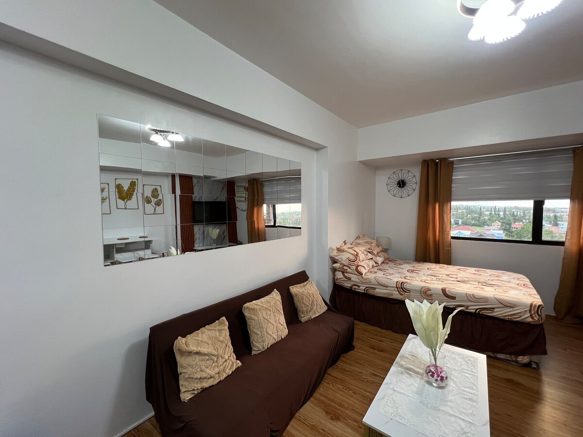 A cozy living area features a brown sofabed accompanied by decorative cushions and a white center table. The double-sized bed is placed against the wall, adorned with patterned bedding. Large windows with light curtains allow natural light to brighten the space, complementing the neutral decor.