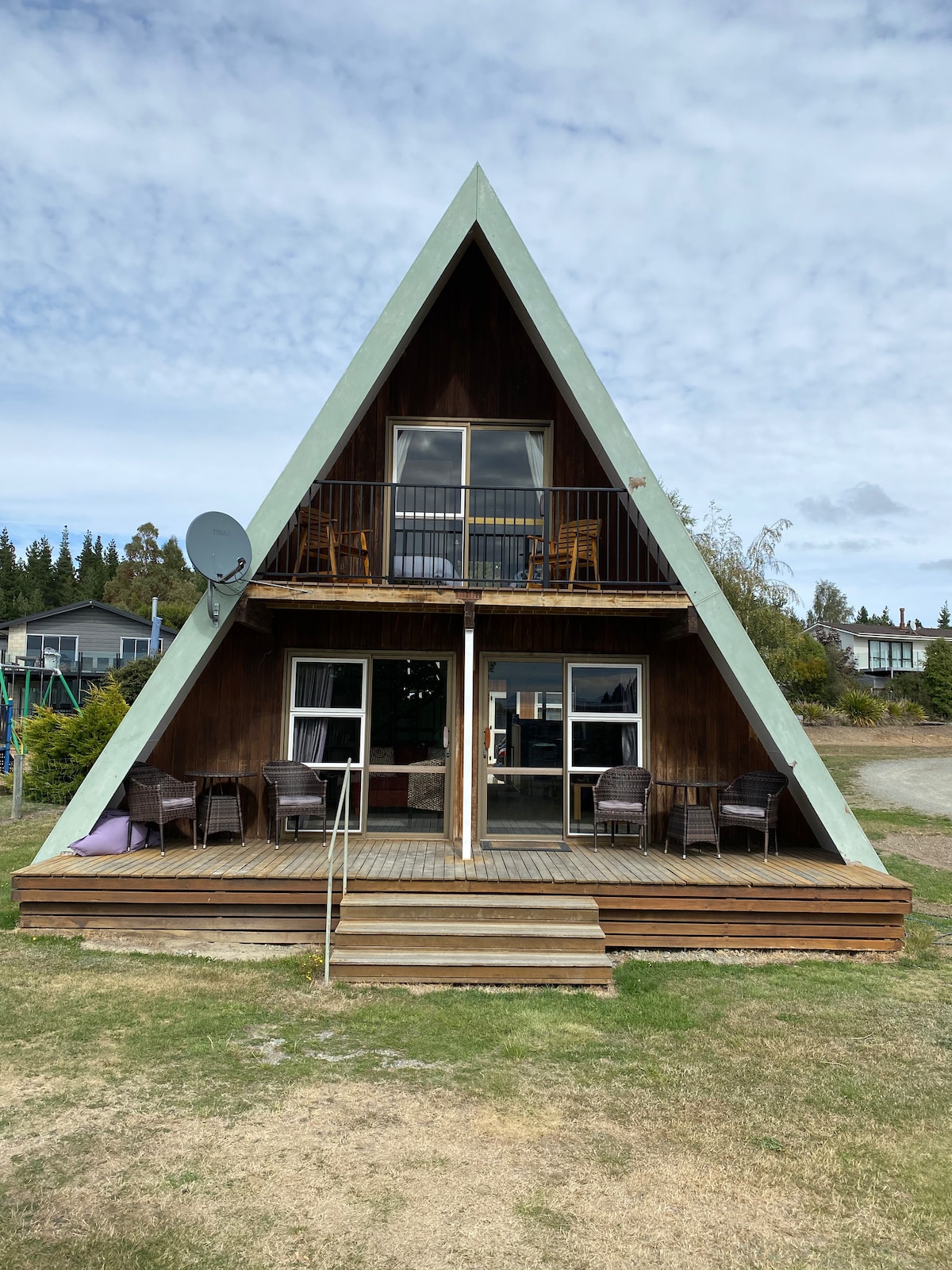 A charming A-frame structure is displayed, featuring wooden siding and a sloping green roof. The exterior is equipped with a spacious deck, adorned with several seating areas and a table, inviting relaxation and social gatherings amidst the natural surroundings.