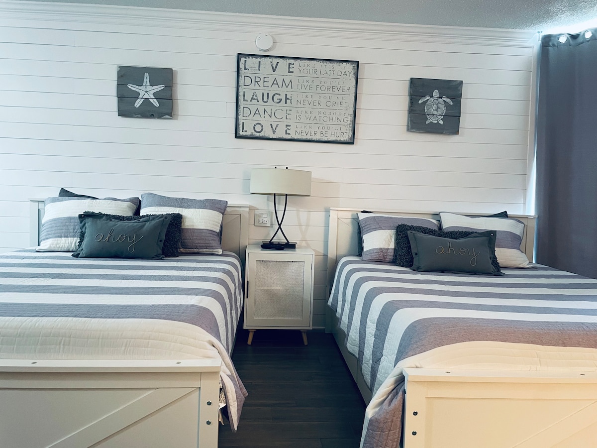 A well-appointed bedroom features two queen beds with striped bedding and decorative pillows. Coastal-themed wall art is displayed above the beds, complementing the light, airy atmosphere. A nightstand is positioned between the beds, adding functionality to the space.