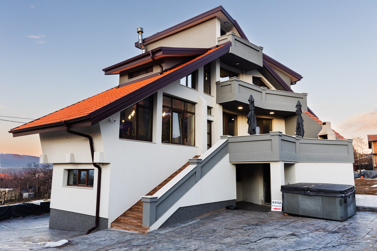 A multi-level house features a distinctive sloping roof with red tiles. Large windows allow natural light to fill the spaces, while a series of stairs provide access to the entryway. Outdoor statues add visual interest to the façade, enhancing the structure’s architectural appeal.