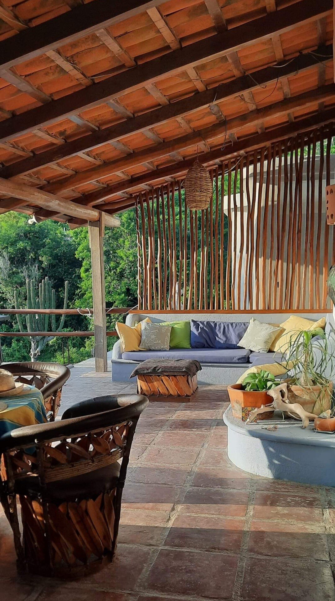 An inviting outdoor seating area features comfortable seating with colorful cushions. A decorative wall made of wooden slats enhances the space, while lush greenery is visible in the background. Natural light illuminates the rustic tiled floor, creating a warm and welcoming environment.